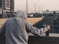 Das Bild zeigt einen auf einer Autobahnbrücke stehenden Mann, der Alkohol in seiner rechten Hand hält